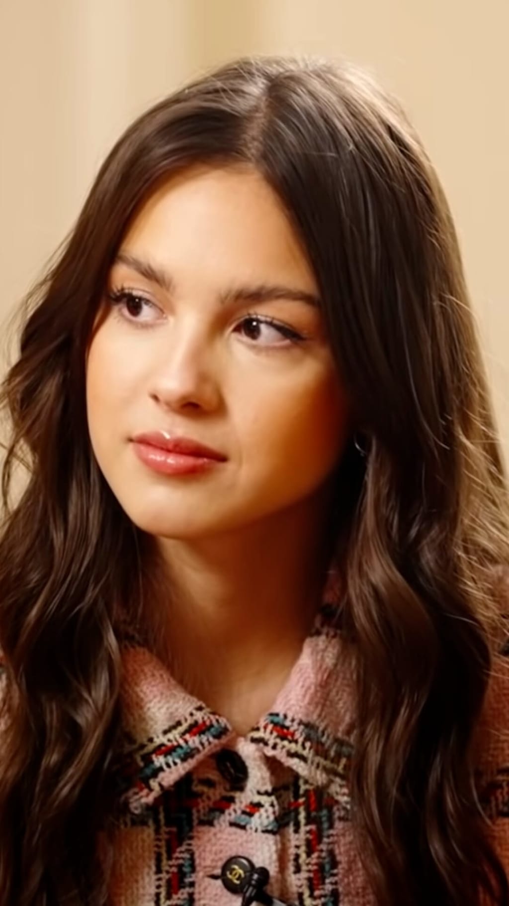 Portrait of Olivia Rodrigo with long dark wavy hair, wearing a patterned sweater, looking downward against a warm beige backg
