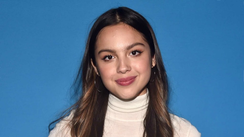Olivia Rodrigo wearing a cream turtleneck sweater against a solid blue background, with long dark hair and subtle makeup