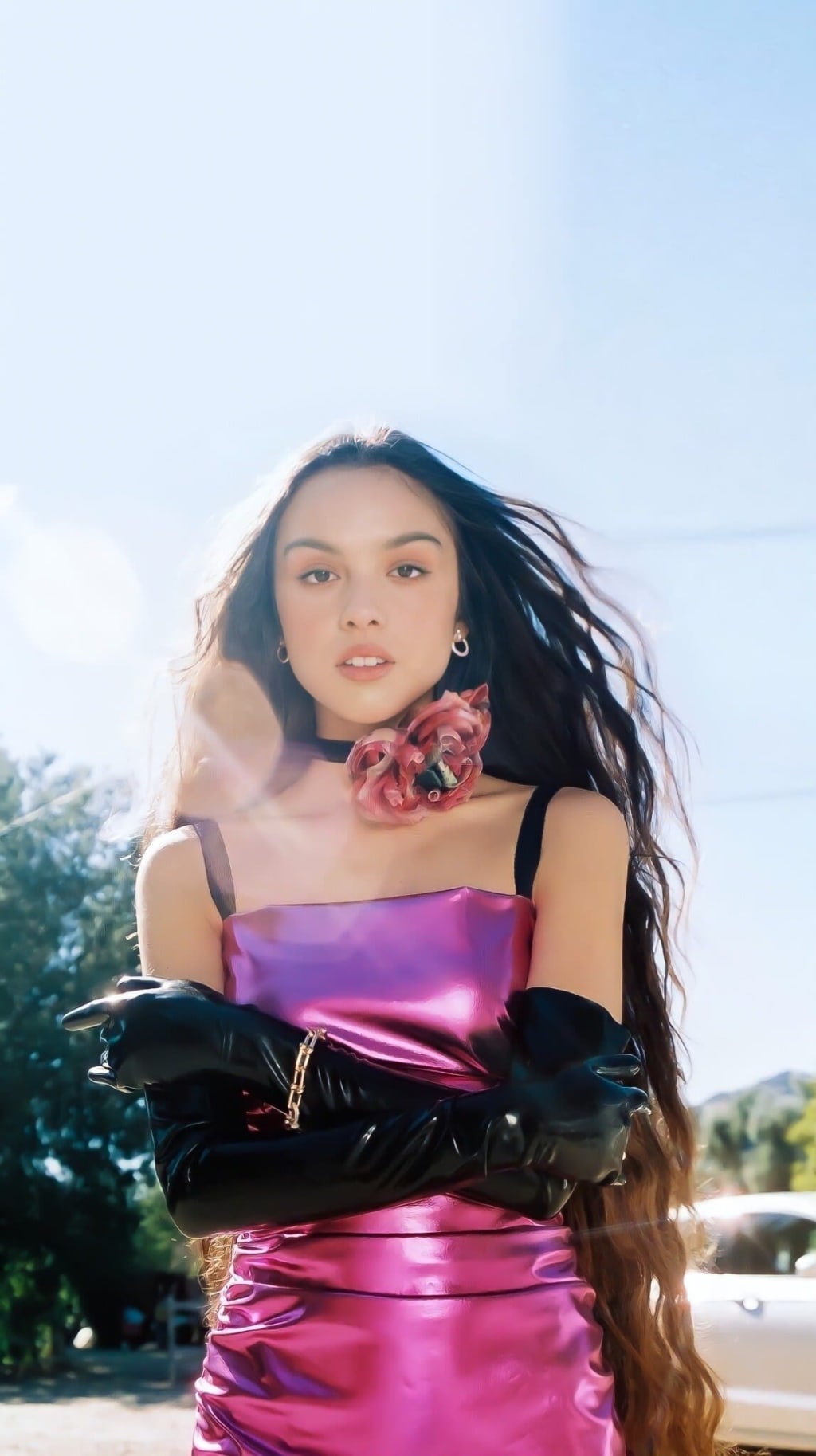 Portrait of Olivia Rodrigo wearing a vibrant pink satin dress with black gloves and pink roses, against a bright outdoor back