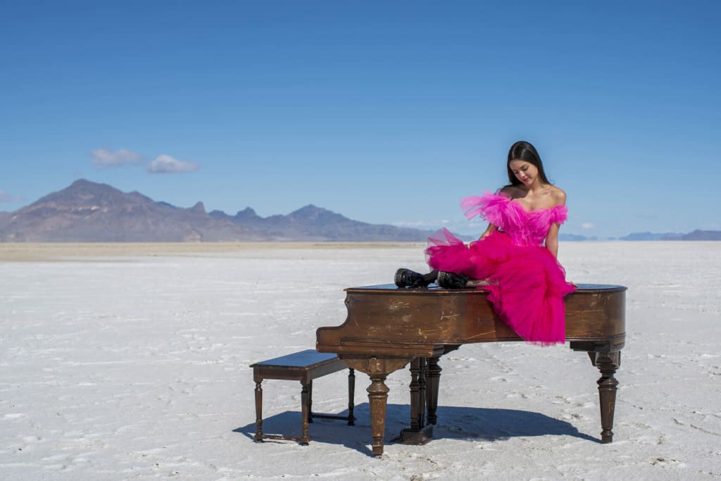 Woman in vibrant pink ruffled dress seated at vintage grand piano on white desert salt flat with mountains in background