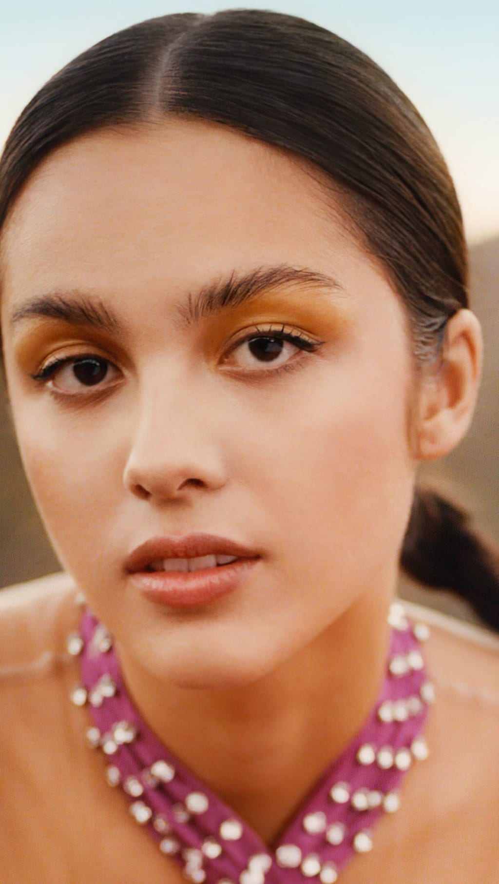Portrait of Olivia Rodrigo with golden eyeshadow, dark straight hair, pink pearl earrings, and neutral expression against sof