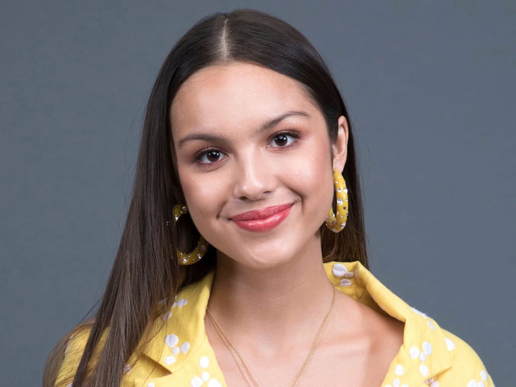 Portrait of a woman with long dark hair wearing a yellow patterned shirt with gold hoop earrings against a gray background
