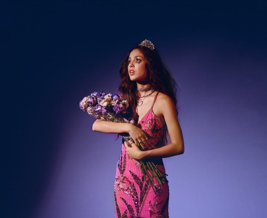 Woman in magenta sequin dress wearing tiara and chain necklace, holding purple and white bouquet against rich purple backgrou