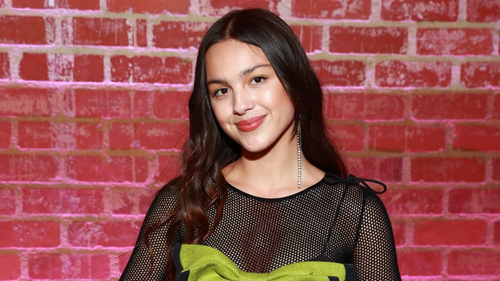 Young woman with long dark hair wearing black mesh top and lime green accent, posed against vibrant red brick wall background