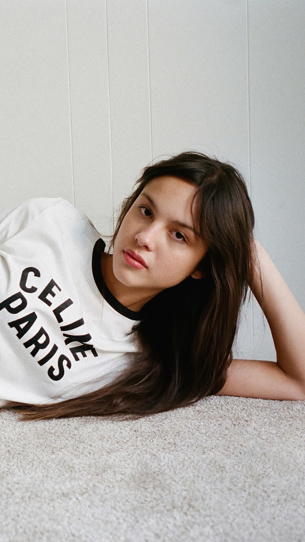 Portrait of a young woman with long dark hair wearing a white graphic t-shirt, posed thoughtfully against a minimalist white
