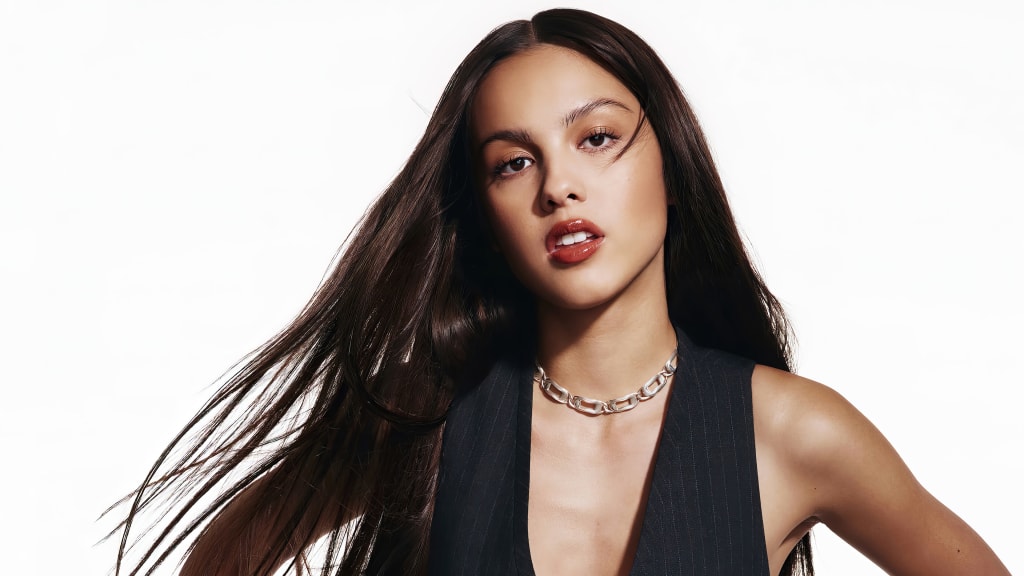 Olivia Rodrigo poses confidently against a white background wearing a black halter top and silver chain necklace, with long d