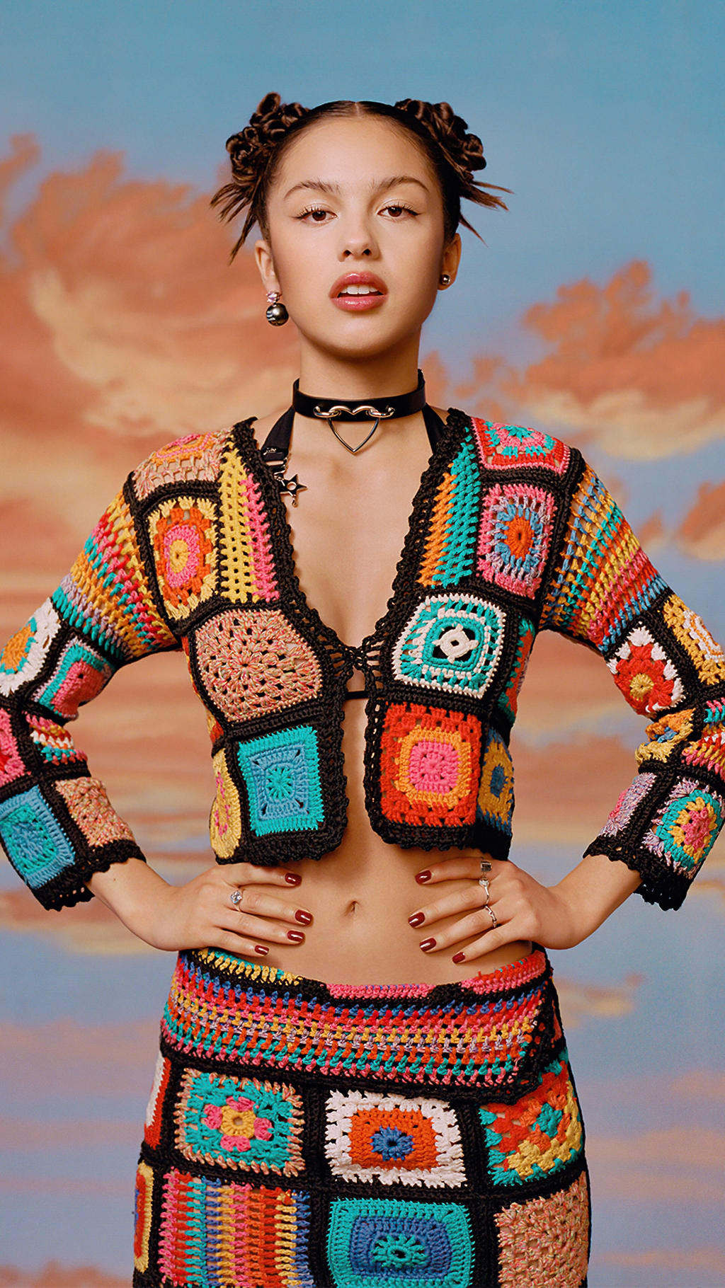 Woman wearing vibrant multicolored crochet crop top and skirt against orange sunset sky backdrop with braided hair and choker