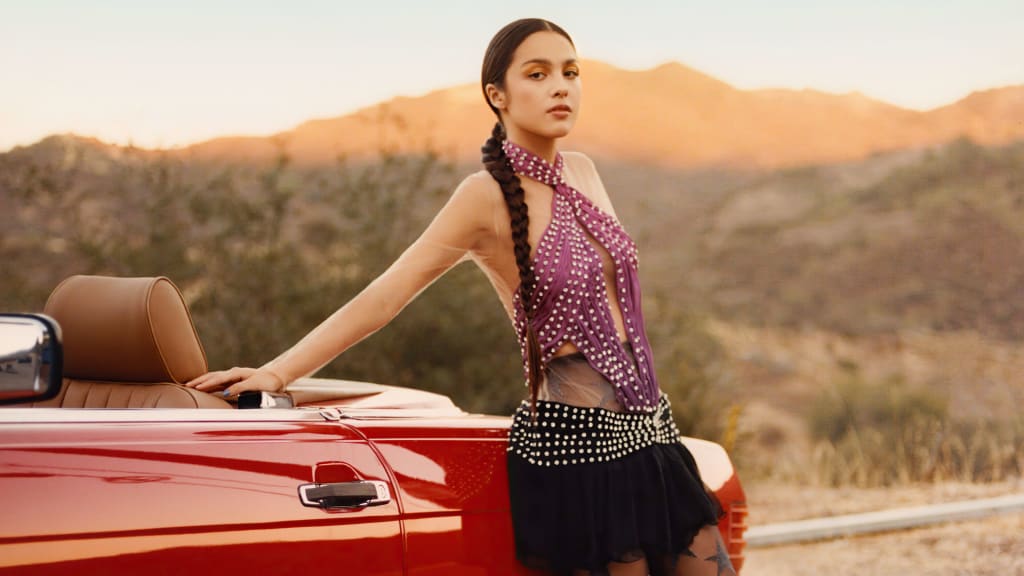 Woman in purple studded outfit leaning against red convertible car with desert mountains and forest landscape at golden hour