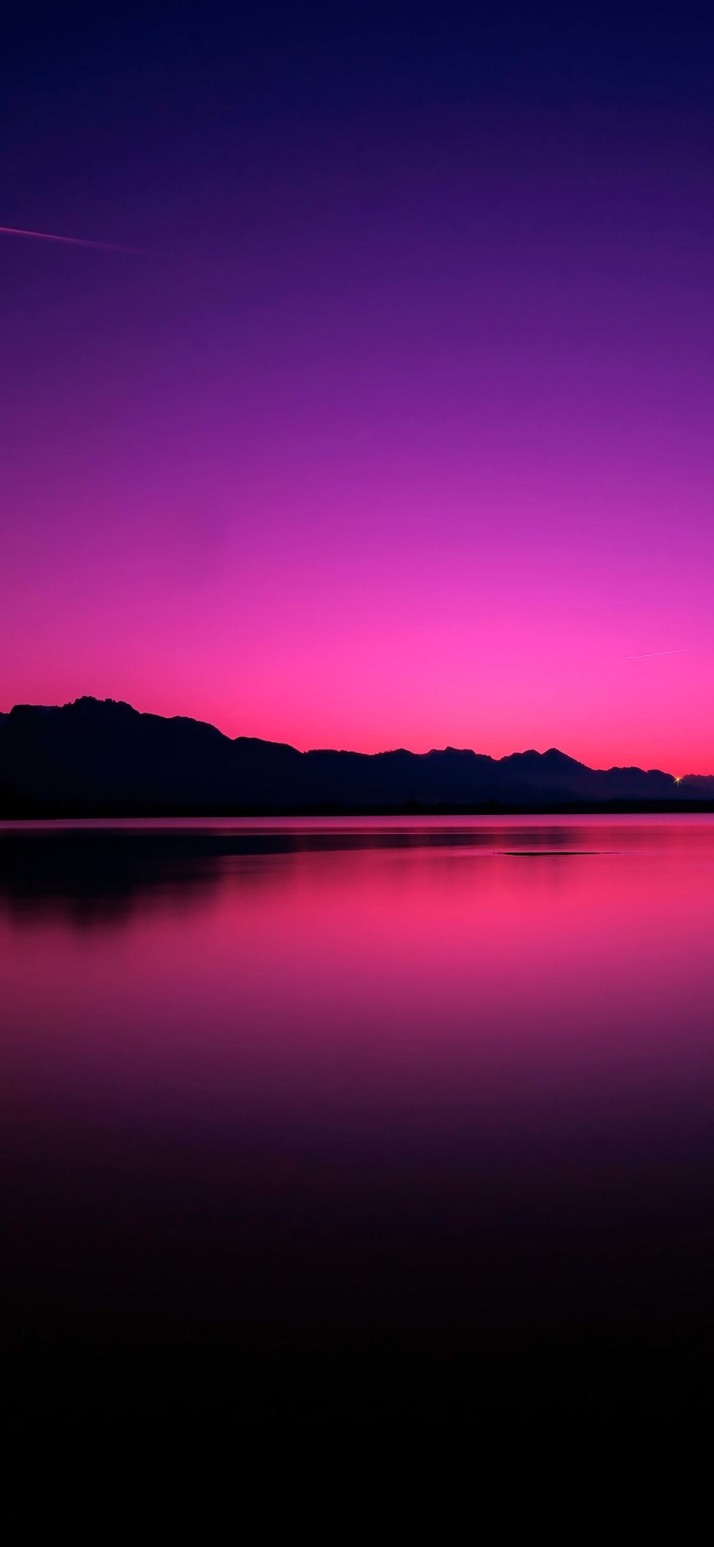Vibrant magenta sunset reflecting on calm lake water with dark mountain silhouettes along horizon
