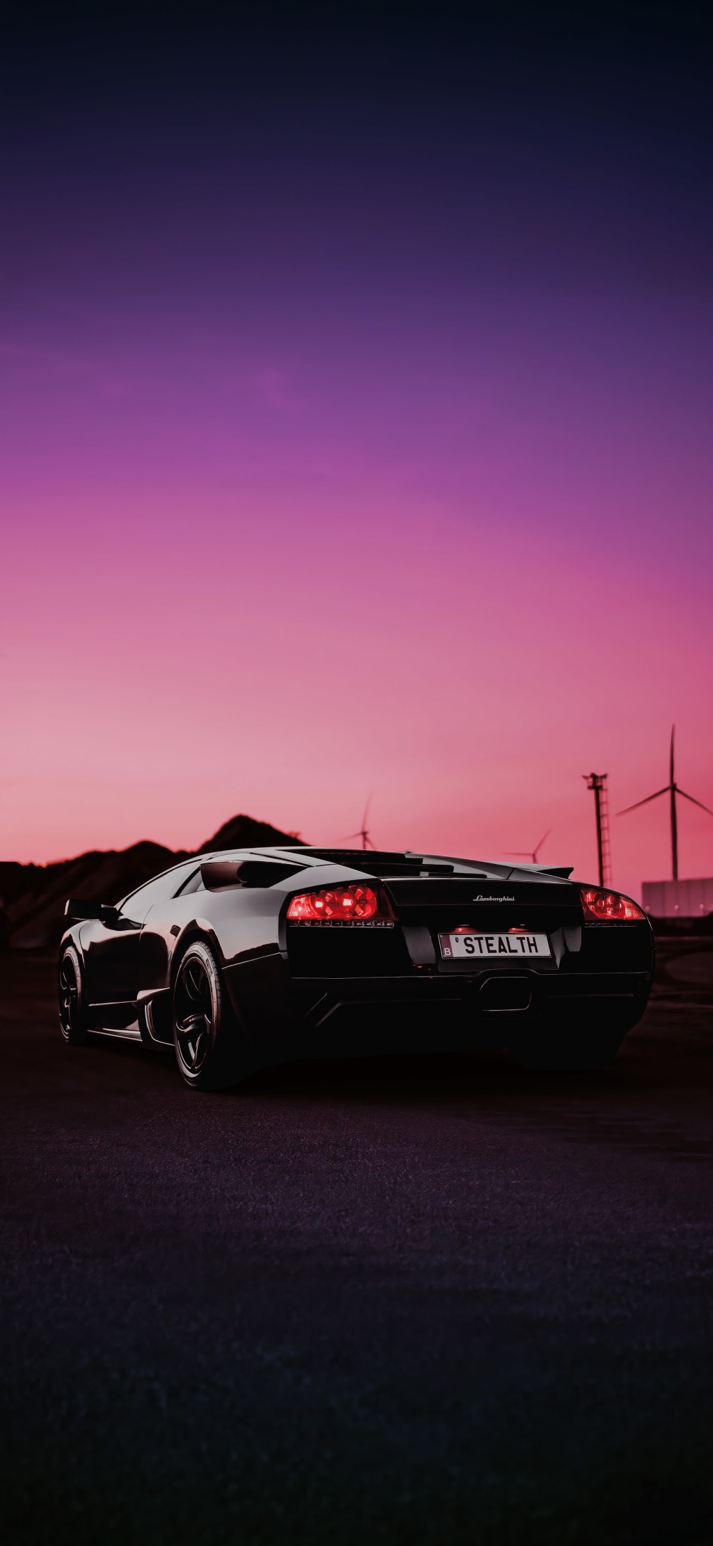 Black luxury supercar parked at dusk with vibrant pink and purple sunset sky, wind turbines silhouetted in background