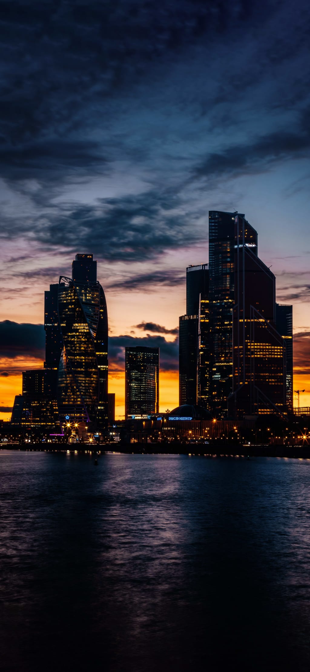 Illuminated city skyscrapers silhouetted against a dramatic sunset sky with golden and blue hues reflecting on calm water bel