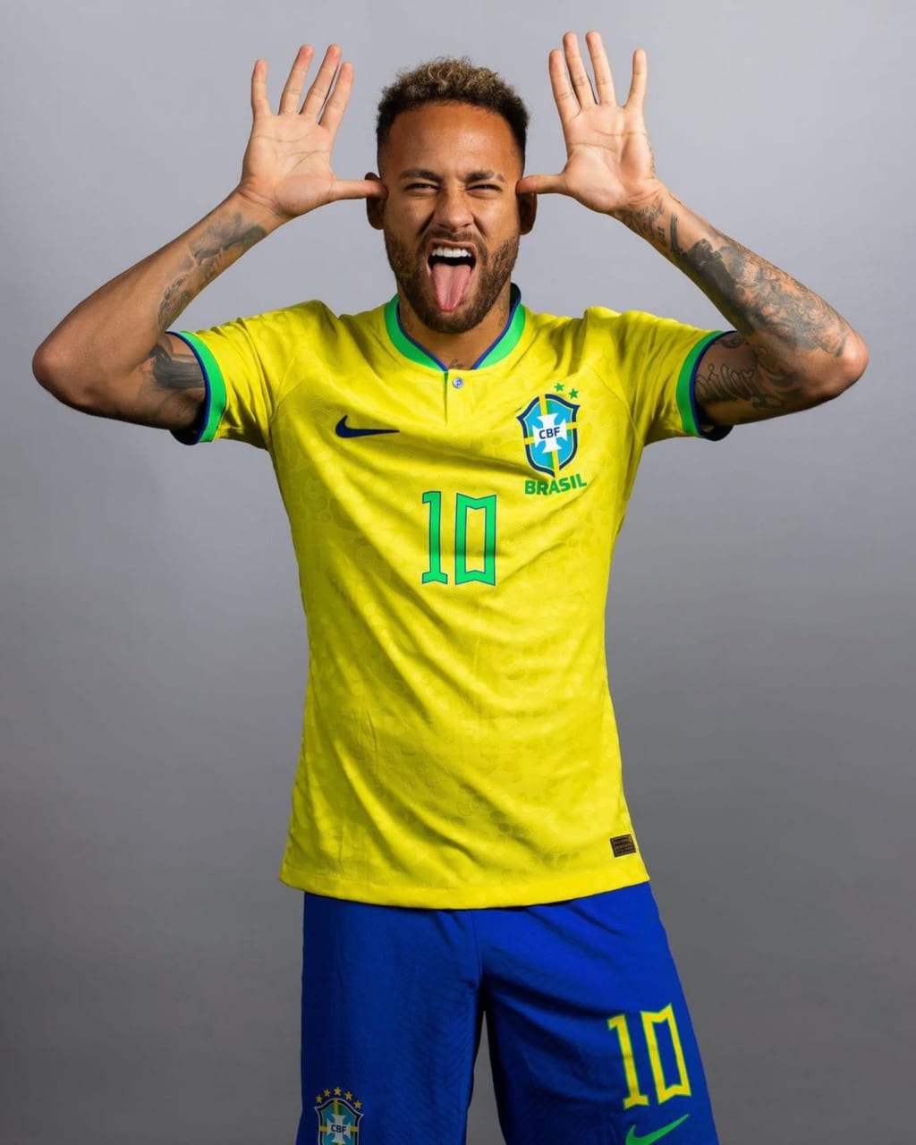 Brazilian footballer Neymar Jr in yellow and blue national team jersey, making a playful face with hands near ears against gr