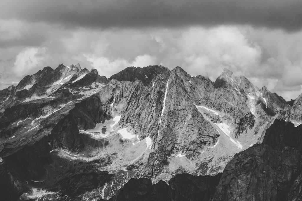 Dramatic black and white mountain landscape with jagged rocky peaks and cascading waterfalls beneath stormy clouds above