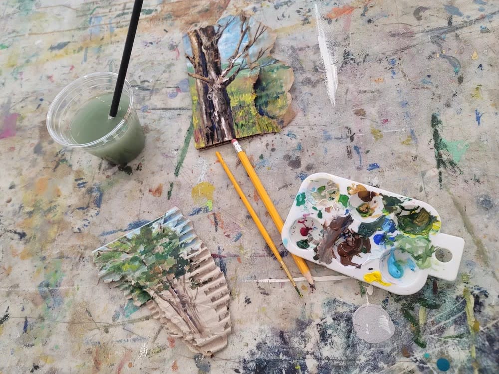 Artist's workspace with painted tree studies, brushes, palette, and water cup on a colorful splattered work surface.