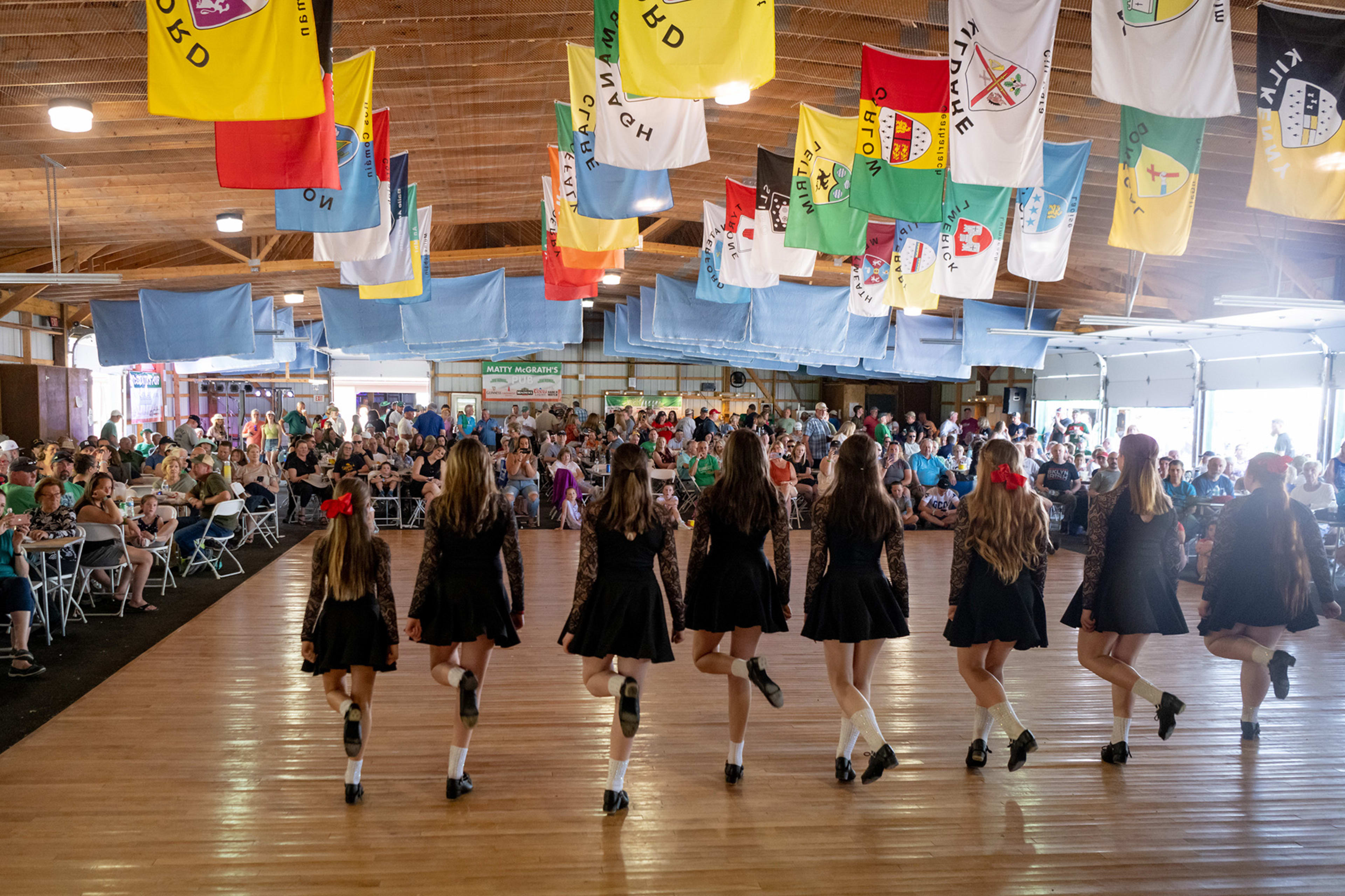 Young dancers in black dresses perform Irish step dancing at an indoor festival hall decorated with colorful flags.