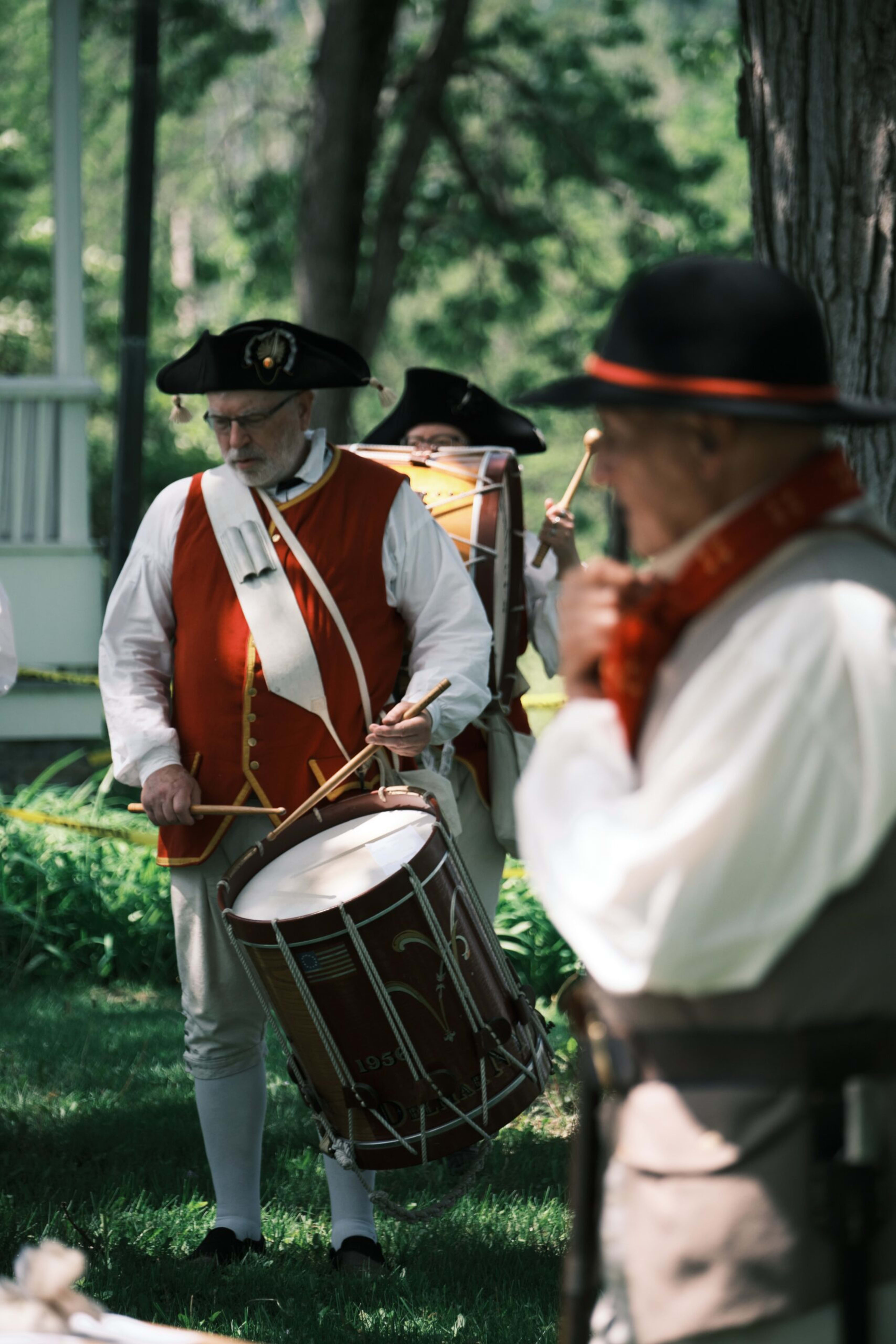 Three men in 18th-century colonial uniforms and tricorn hats perform with drums outdoors among trees.