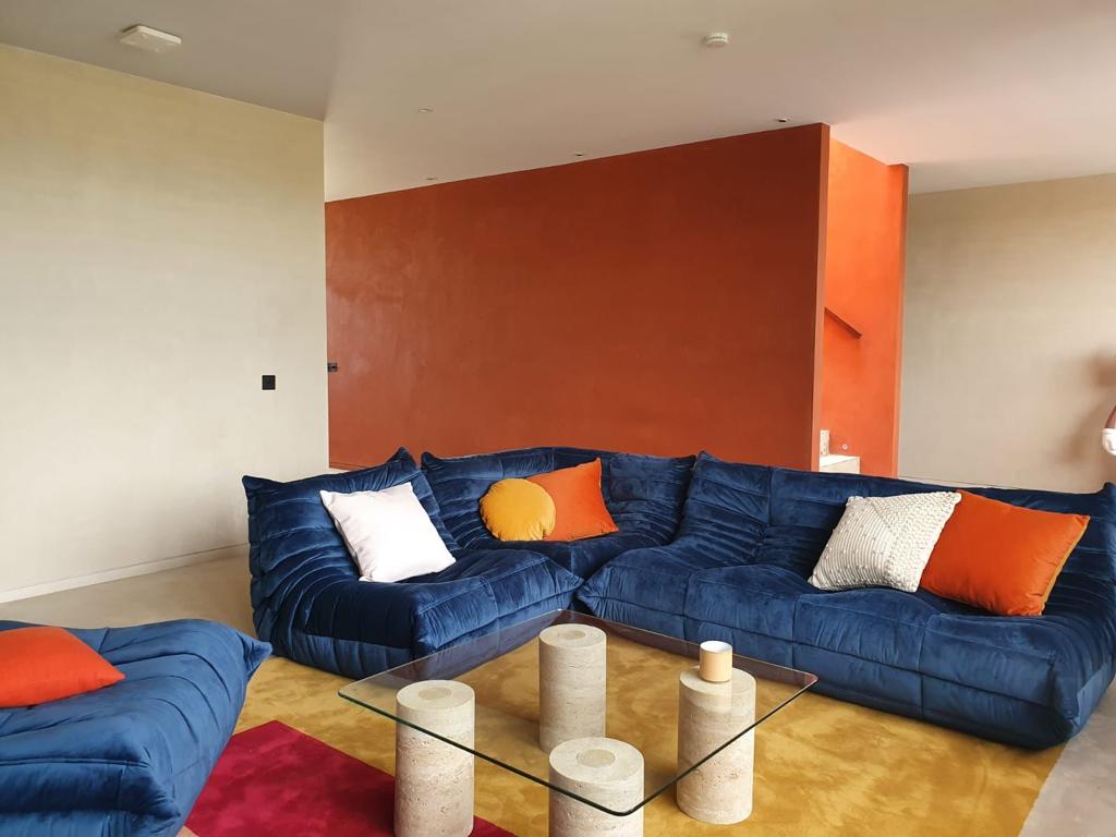 A royal blue Togo corner sofa with orange and white cushions against a terracotta wall, on a yellow rug with a glass and travertine table