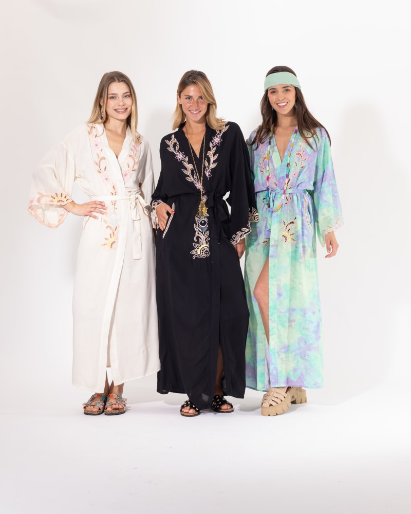 Trois femmes souriantes portant des kimonos longs bohèmes à broderies florales et motifs ajourés, déclinés en trois coloris : écru, noir et tie-dye vert/violet, ceinturés à la taille.
