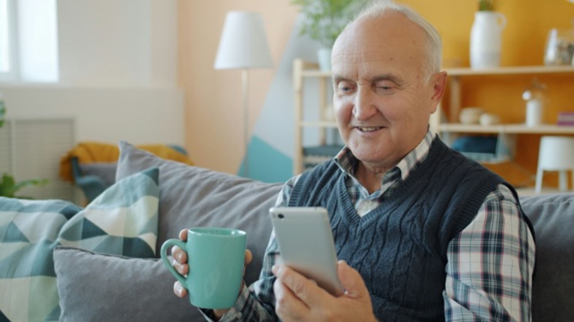Homme senior souriant tenant une tasse et un téléphone assis sur un canapé.