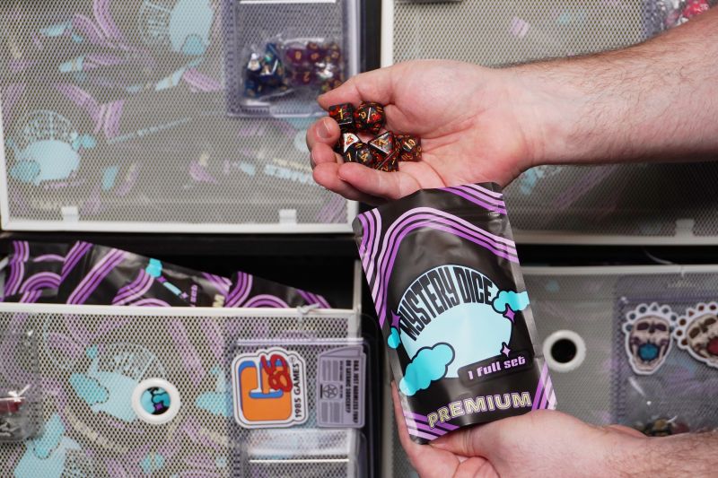 Hand pouring colorful polyhedral dice set into Premium Mystery blind bag.
