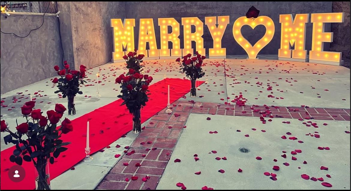 Romantic Marry Me marquee letter proposal setup with rose petals and candles by Sassy Decorations