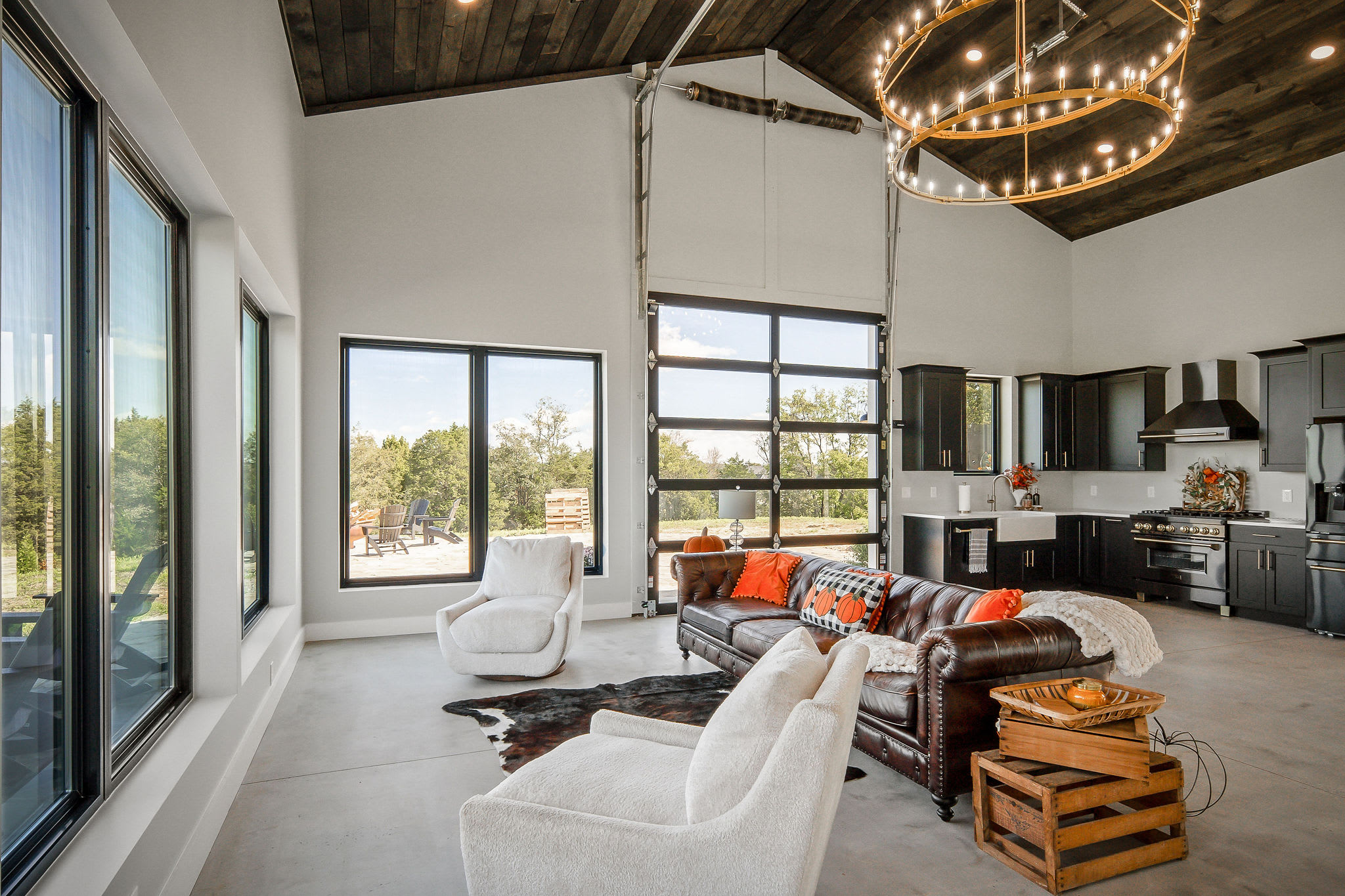 Open interior of the guest home and event space with a brown leather Chesterfield sofa, white swivel chairs, twin candle chandeliers, a dark stained vaulted plank ceiling, and a glass roll-up garage door opening to the patio.