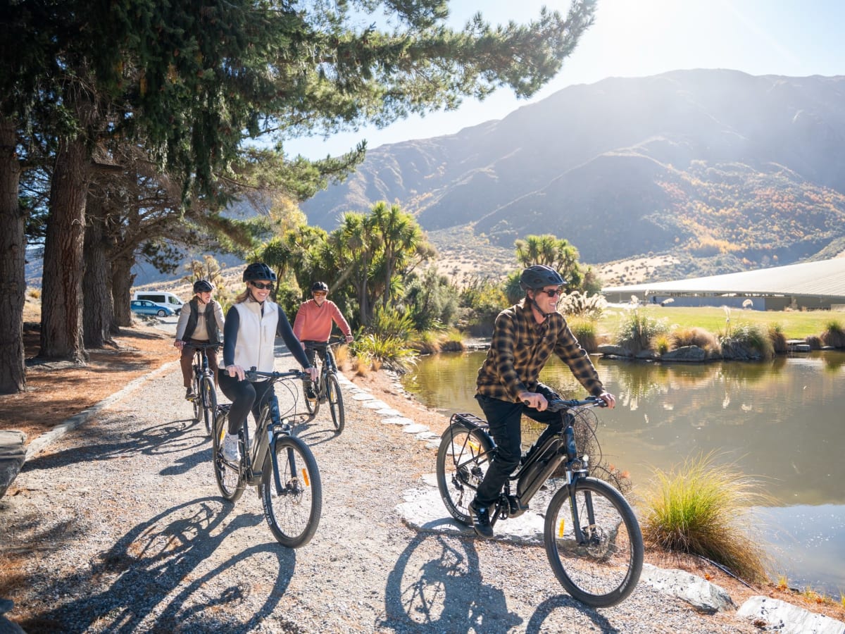 Group of e-bike riders cresting a Gibbston Valley vineyard with mountains and a pond behind them