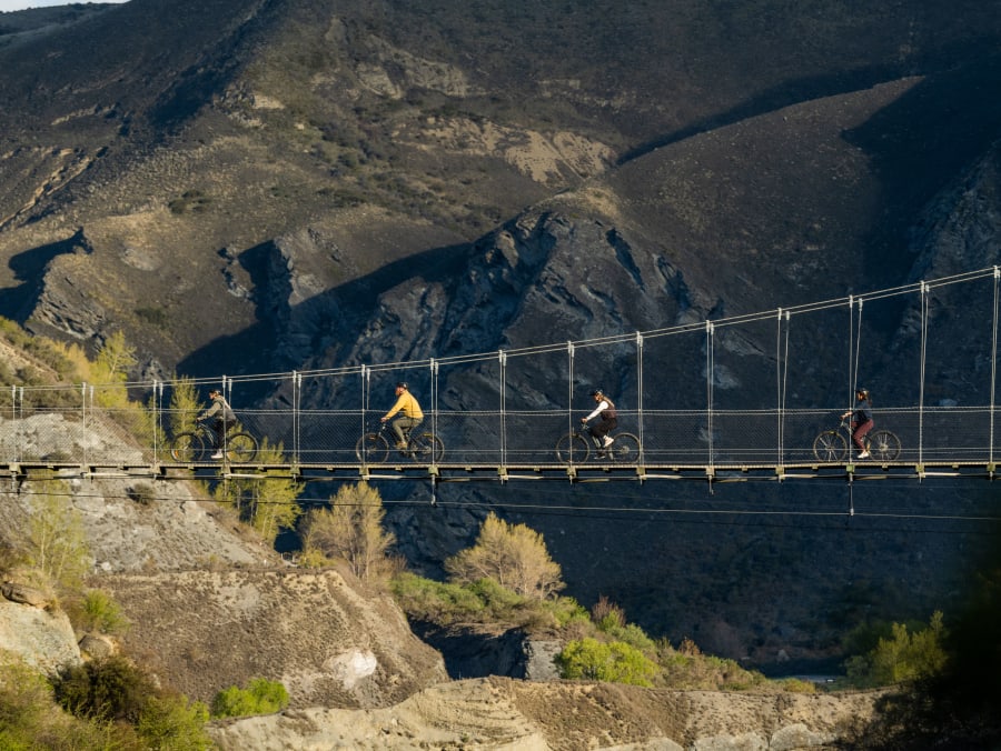 E-bike riders crossing a suspension bridge over the Arrow Gorge with rugged schist canyon walls behind