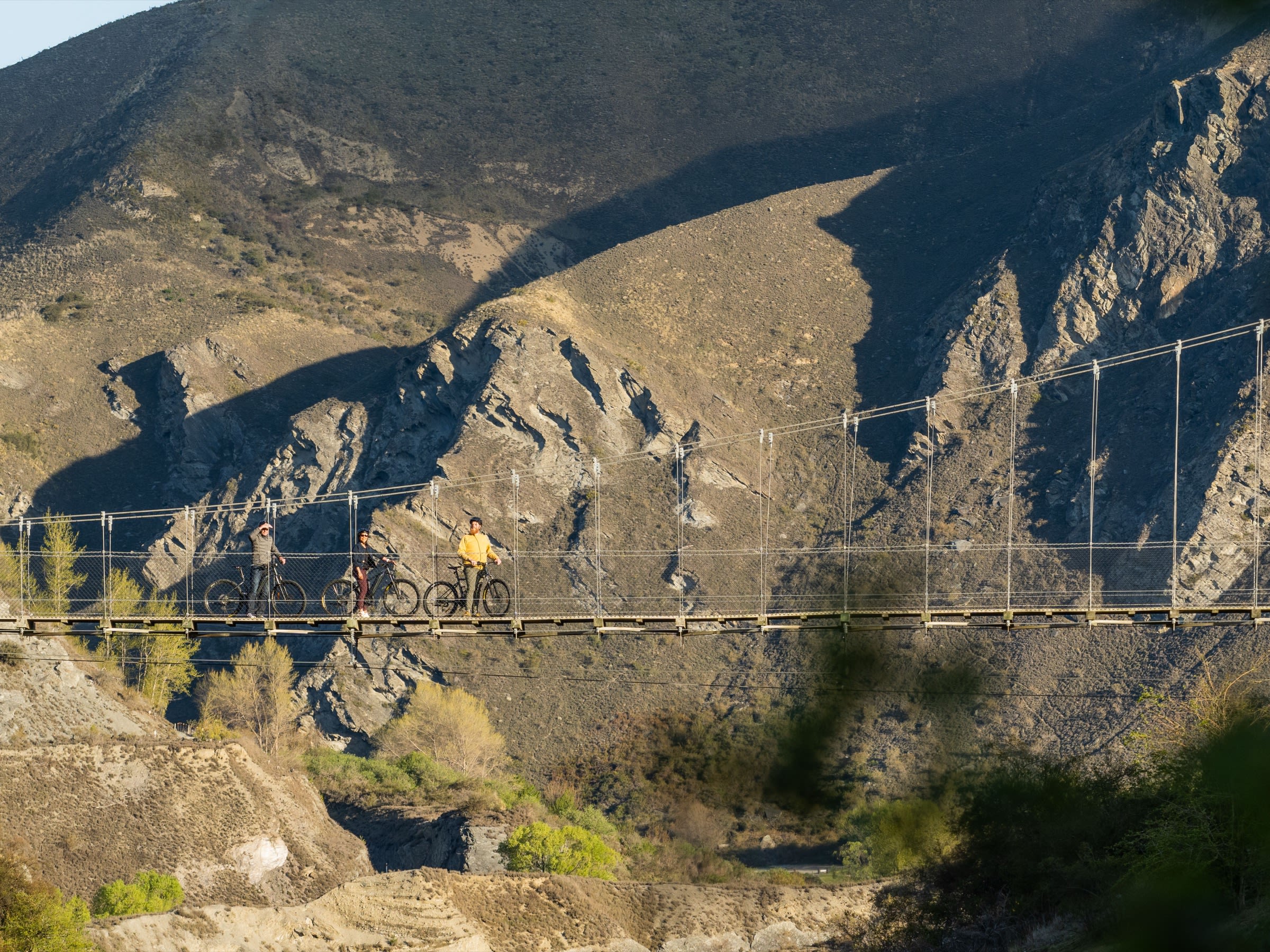 Riders standing at the centre of the 80-metre Edgar Suspension Bridge spanning the Arrow Gorge with rugged schist walls behind