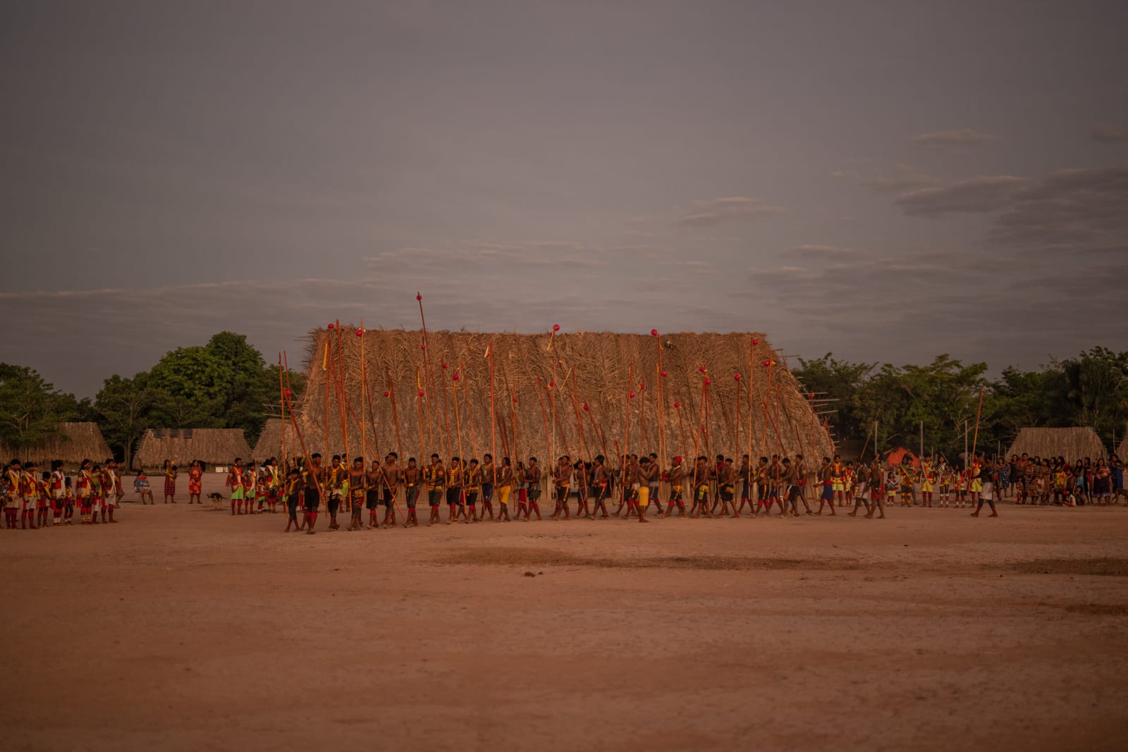 Mebengokre naming festival at dusk, rows of people holding tall ceremonial poles