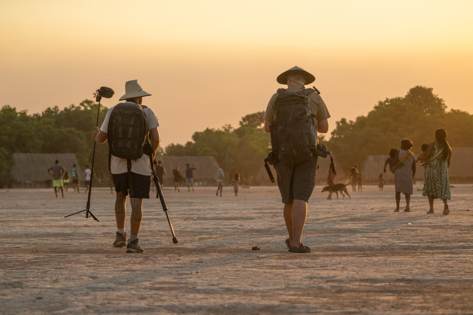 Two filmmakers walking through a village at sunset carrying camera gear and a boom stand