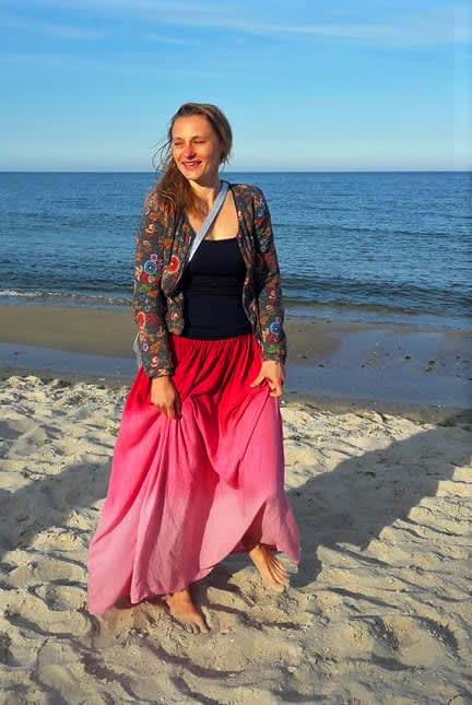 Kobieta w długiej, różowej spódnicy stoi na plaży, uśmiechając się, z morzem w tle. Idealna sceneria na wakacje z jogą.
