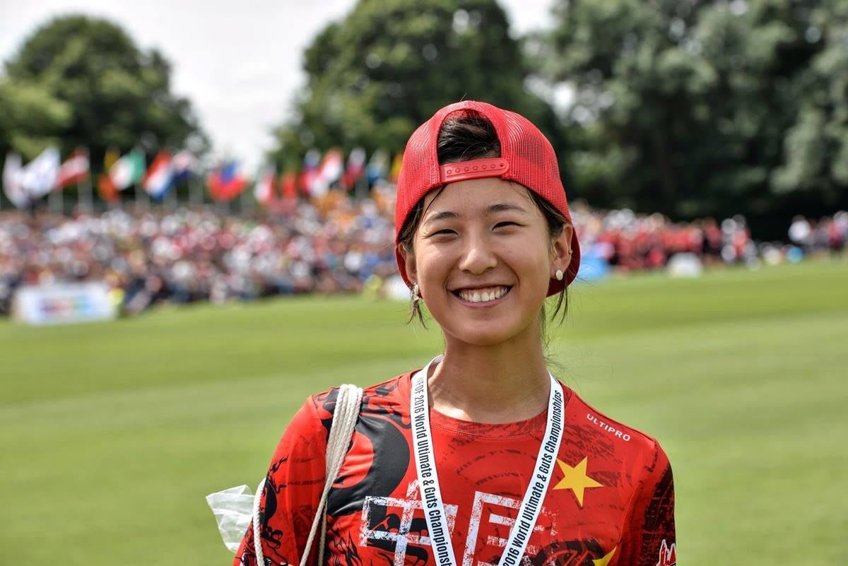 A Chinese female frisbee player smiles at the camera with your fed hat flipped back and her medal around her neck.