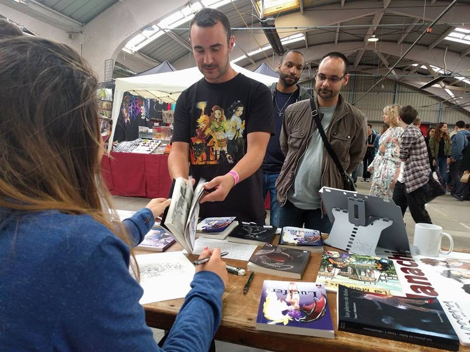 A man hands Lina a book across a table for her to sign as others wait behind him. fans, patience, hard work.