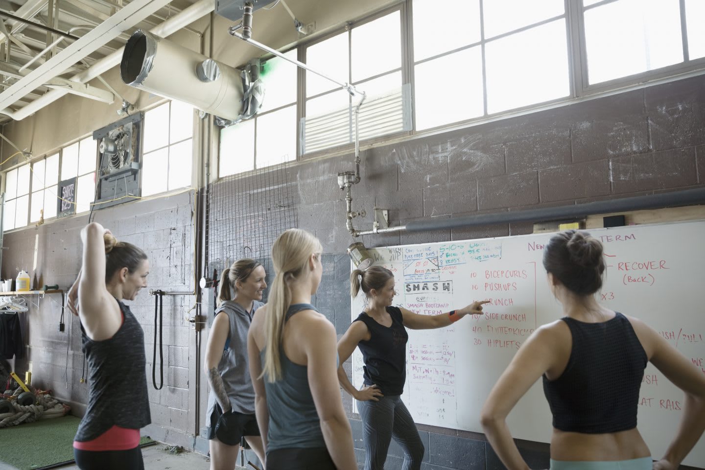 A female CrossFit instructor stands in front of a white board planning a class with her female students in a gritty gym. goal-setting, success, GamePlan A, confidence