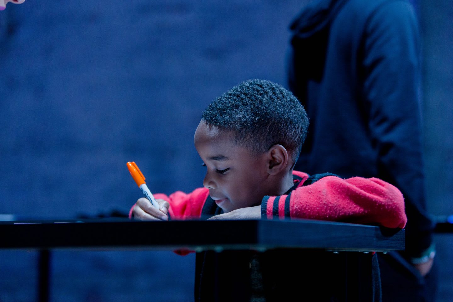 A little boy leans on a table as he draws on a piece of paper with a pen. creative, imagination, creativity, drawing