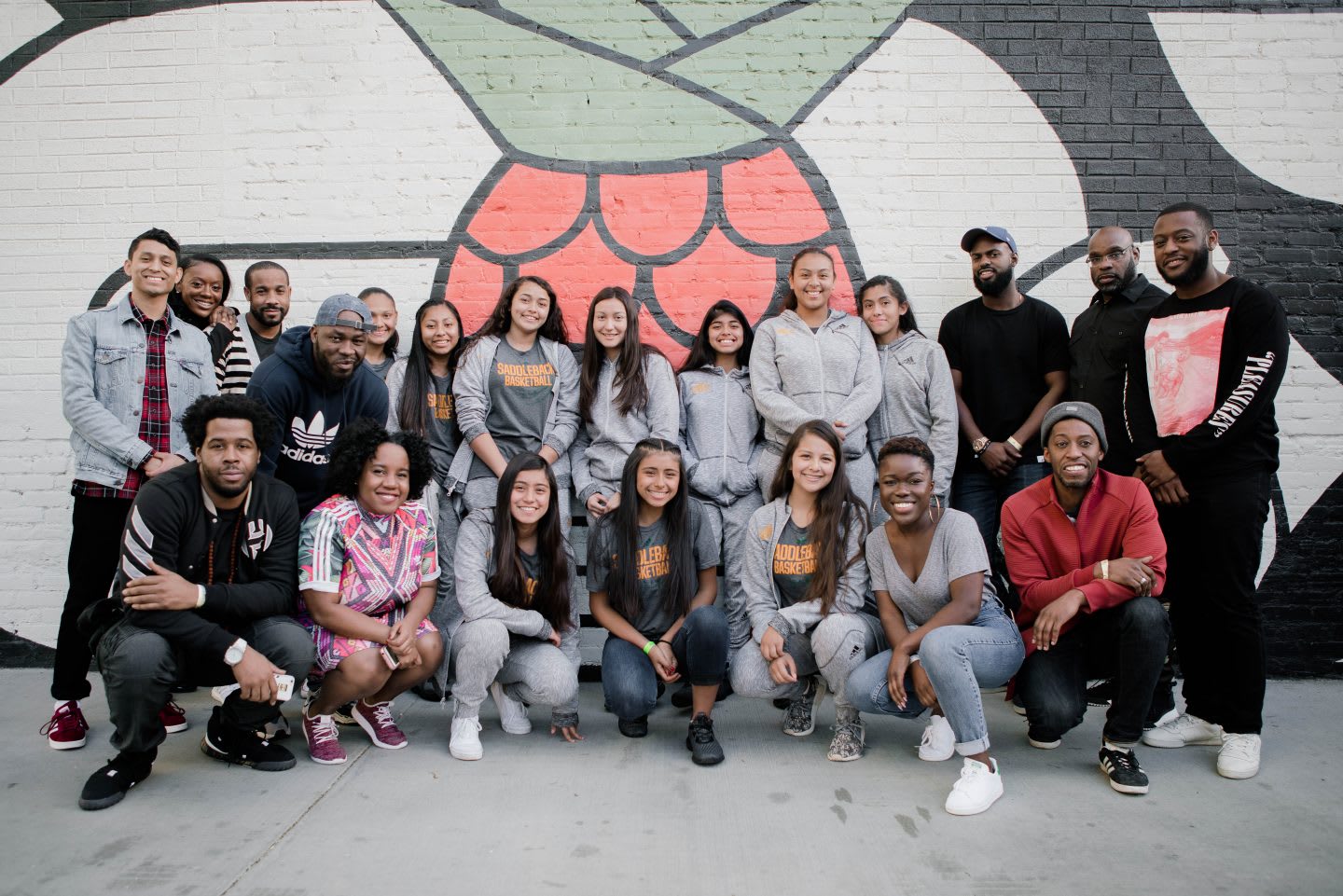A group of youth and mentors pose for a group photo in front of a wall with a mural on it. team, teamwork, celebration, pride