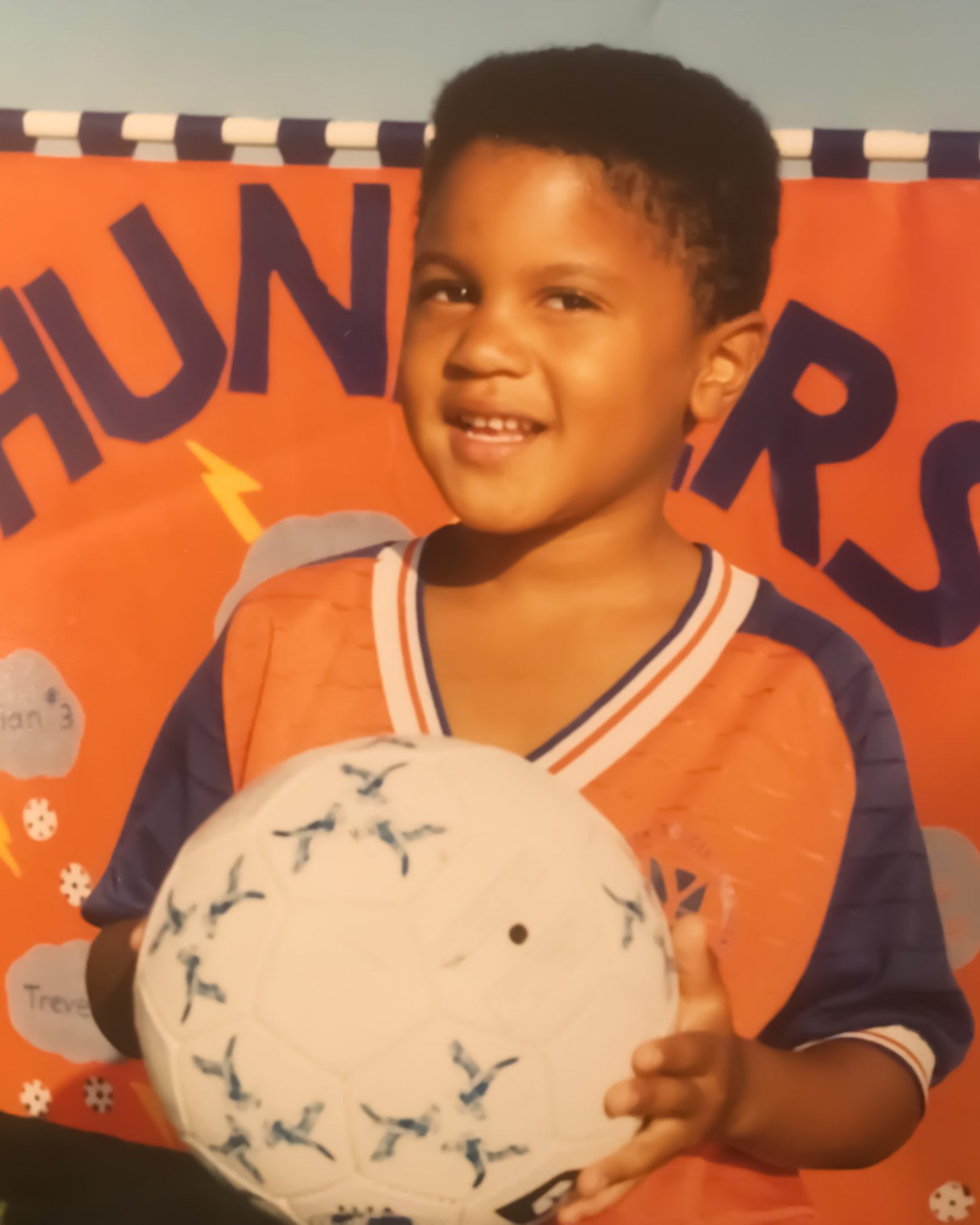 A child holds a football wearing his favorite football jersey. child, football, soccer, ball, jersey