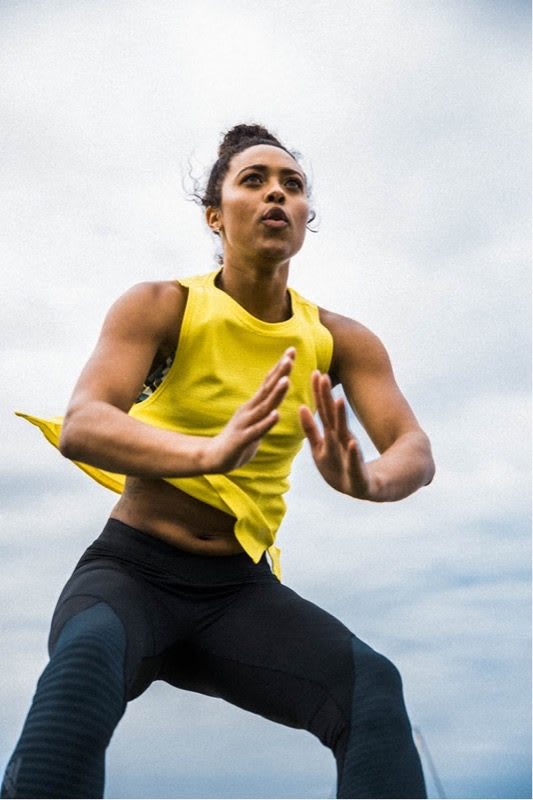 Woman wearing a yellow tank top and black tights is doing a jump squat. Fitness-Jump Squats-Workout-Woman