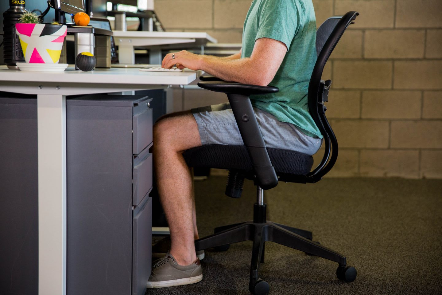 A man sitting at a desk in a healthy sitting position wearing shorts, t-shirt and sneakers. sitting position, health, healthy work place, work space, mental health, productivity, EXOS, GamePlan A