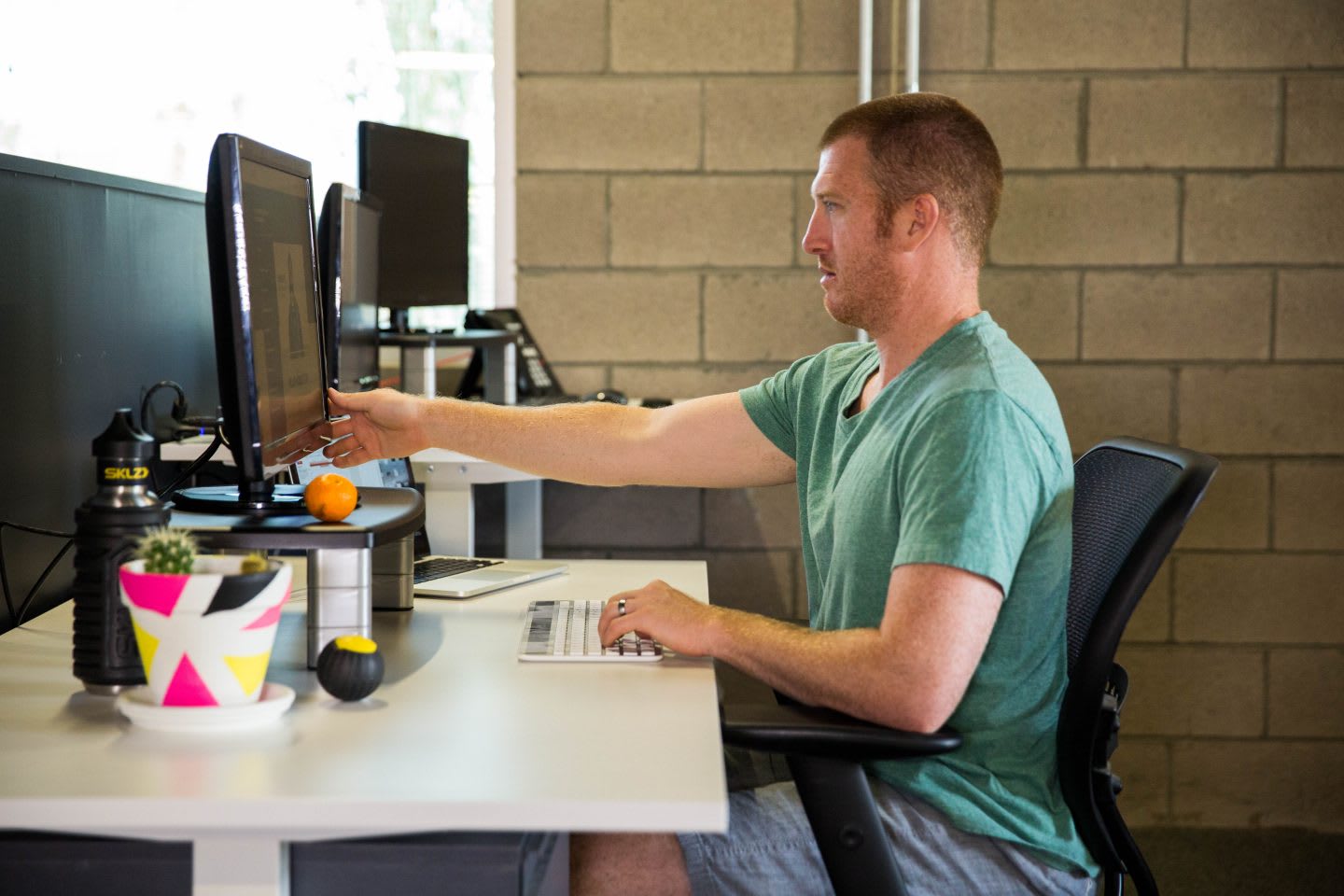 A man sitting at a desk in a healthy sitting position wearing shorts, t-shirt and sneakers. sitting position, health, healthy work place, work space, mental health, productivity, EXOS, GamePlan A