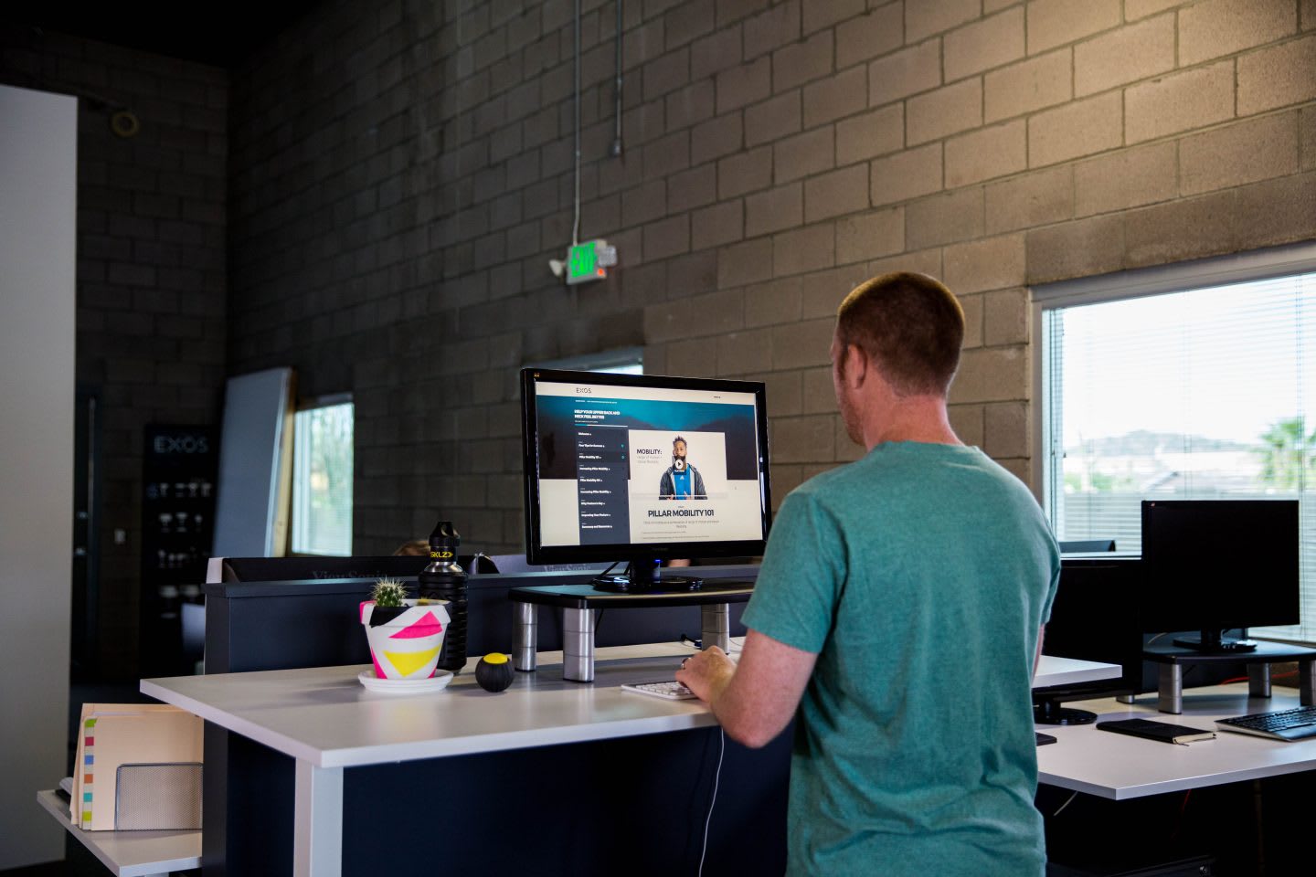 A man standing at a desk in a healthy position wearing t-shirt. standing desk, health, healthy work place, work space, mental health, productivity, EXOS, GamePlan A