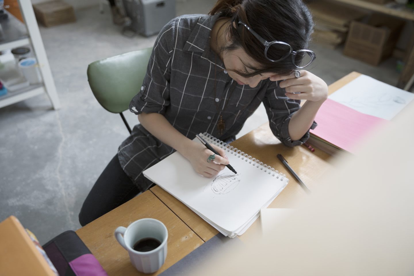 Woman with sunglasses sketching at a desk with concentration, studio, artist, female, coffee cup, notebook, art, focus, creativity, GamePlan A