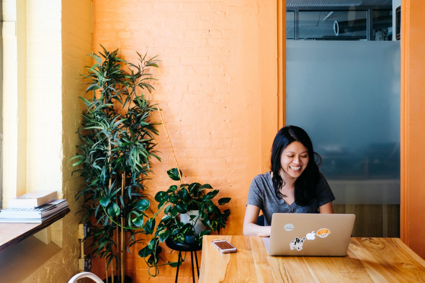 Smiling woman working in cafe with orange walls with laptop surrounded by plants, Georgette Eva, working, happy, inspiration, productivity, new spaces, co-working, GamePlan A