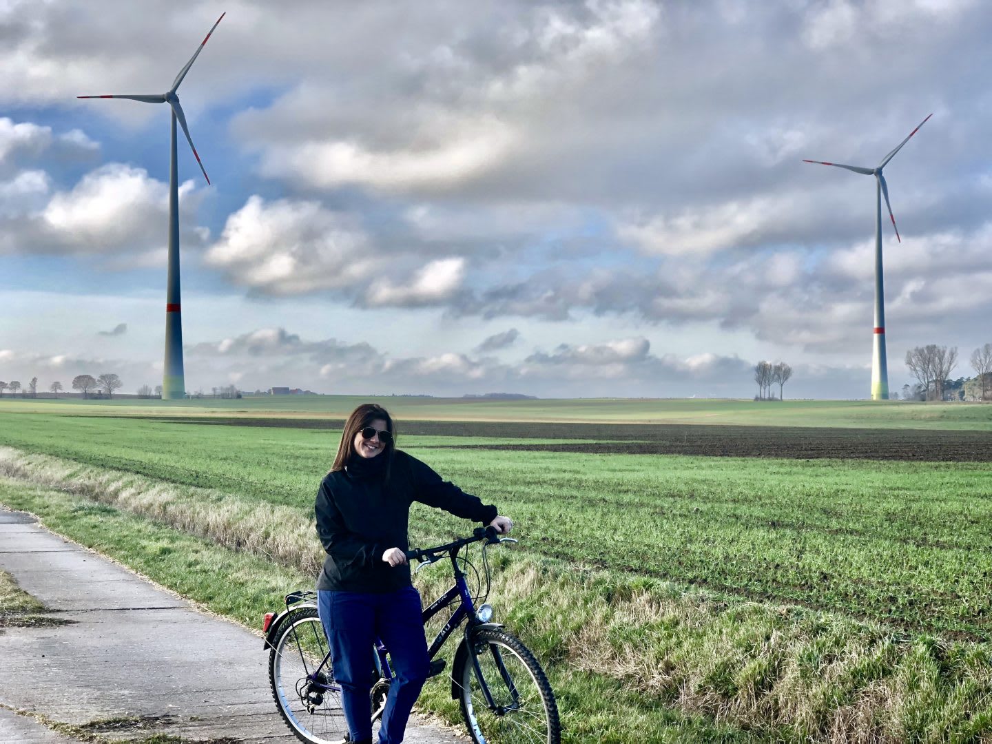 A person standing on a cycling track with her bike in front of wind wheels. sustainability, cycling to work, commuting, GamePlan A