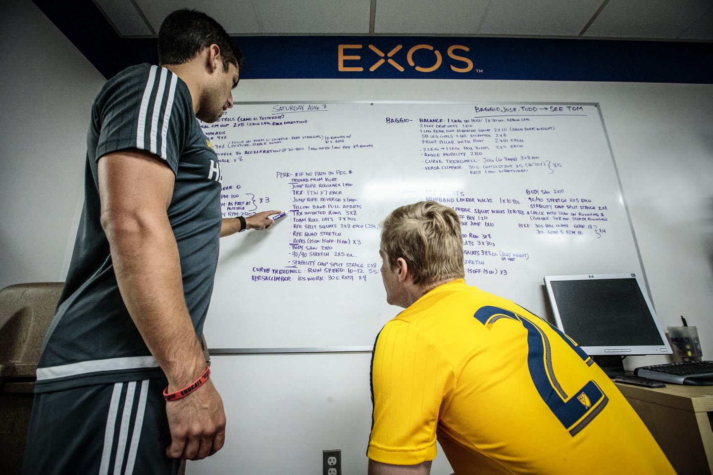 Two men in gym clothes sitting in front of a white board discussing steps of a strategy. goal setting, planning, consulting, achieving goals, strategizing, GamePlan A