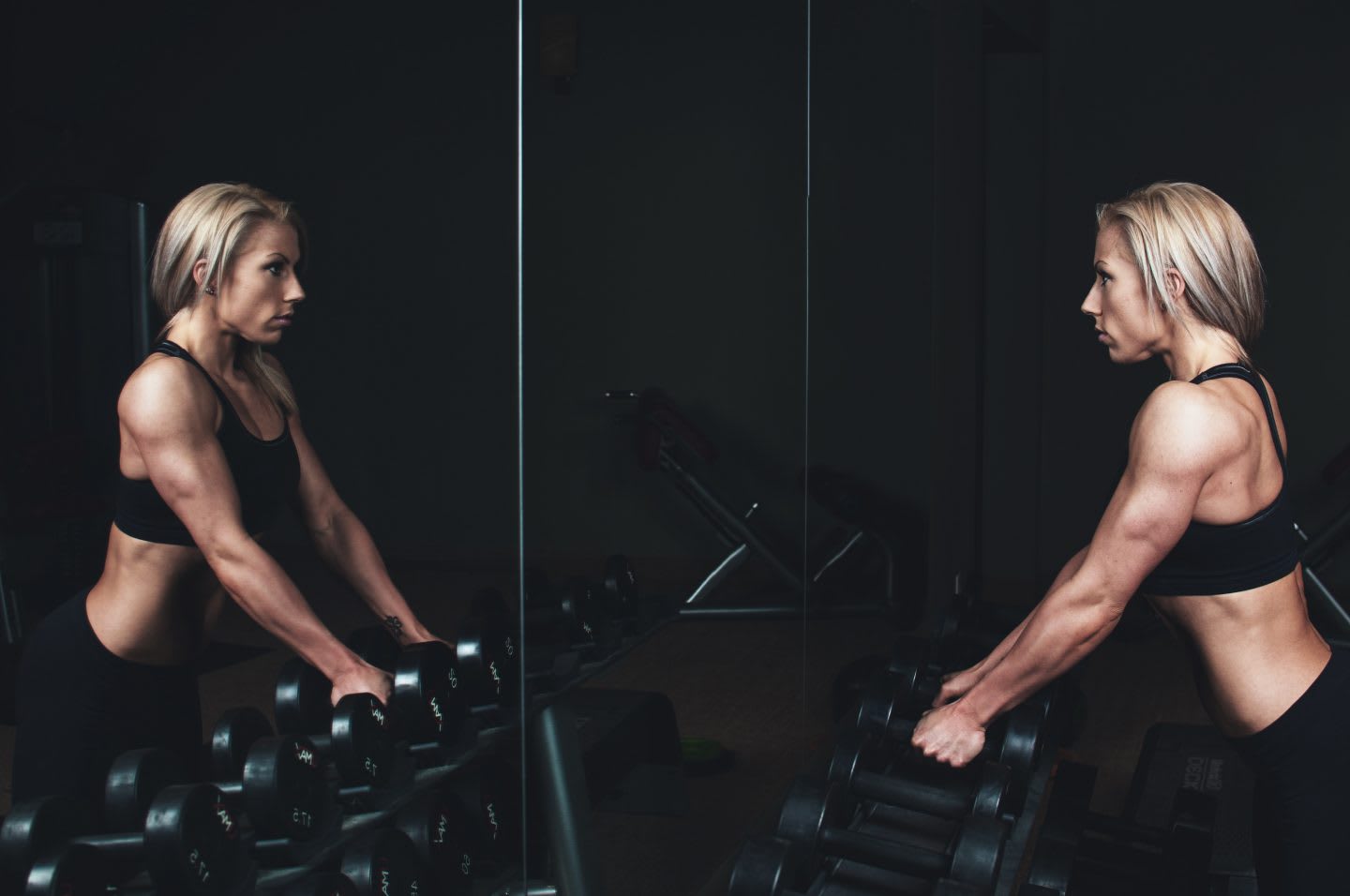 A strong woman looking at her reflection in the mirror in a dark gym, holding weights. self-reflection, self-awareness, body shape, fitness, GamePlan A.