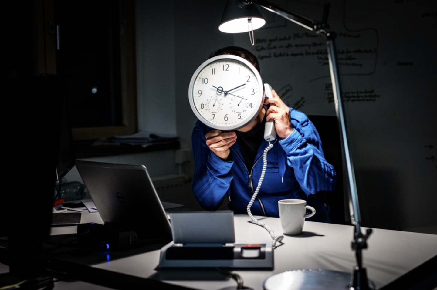 An employee sitting at his office desk late at night, talking on the phone, with a big clock in front of his face. over hours, time management, work-life-balance, performance, GamePlan A