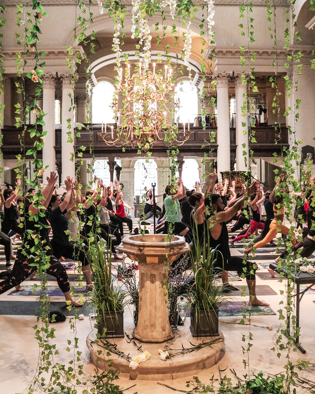 In a beautiful church a cascade of green vines hangs in front of an ornate font, whilst people do yoga in the background. Wellnerss, Gameplan A.