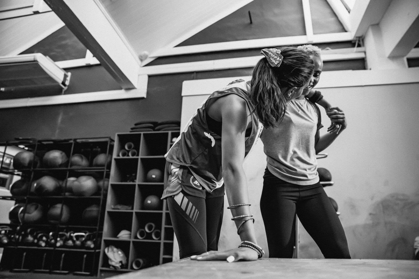 Two women standing in a gym room, one leaning on the other.